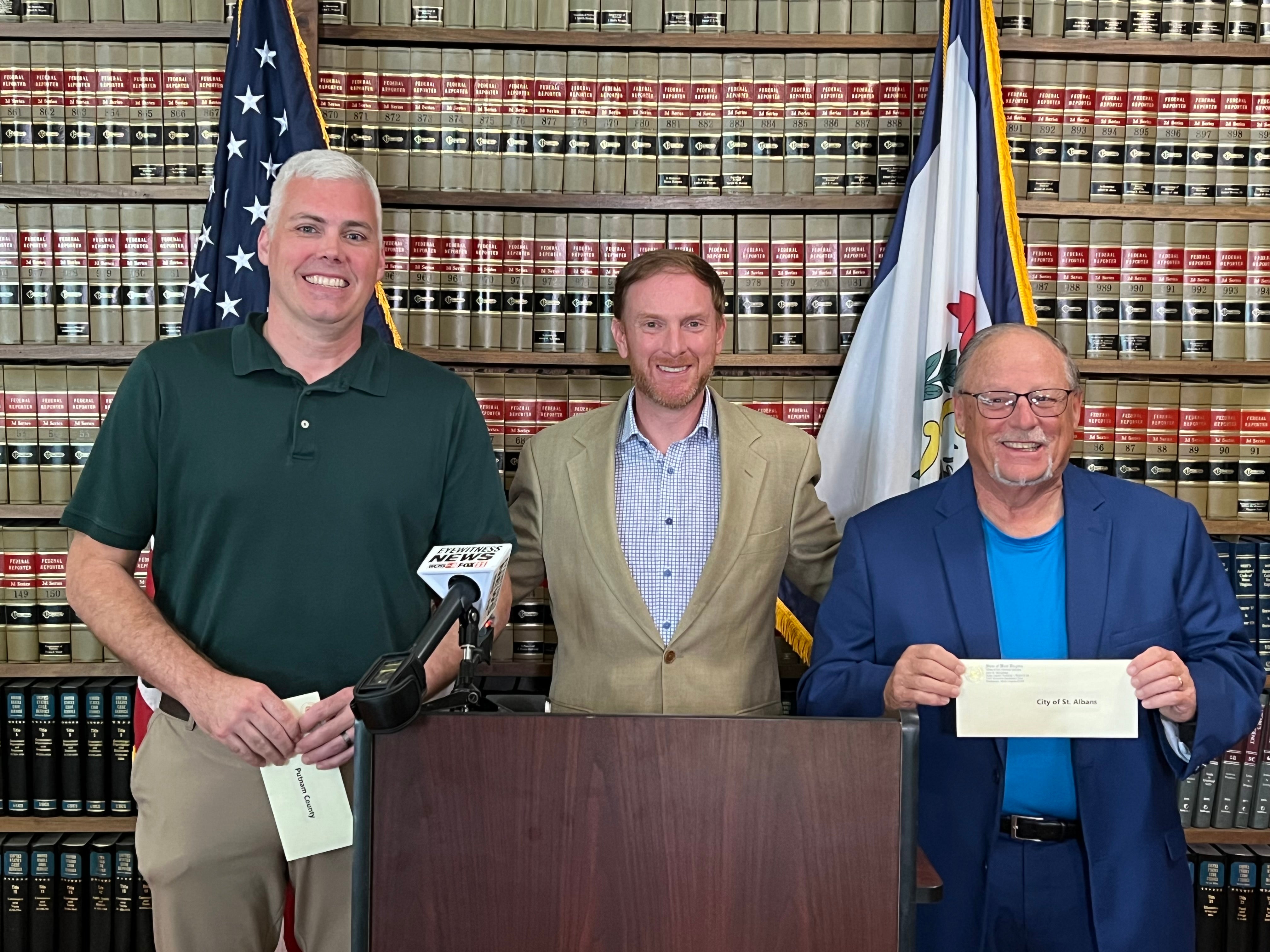 Attorney General JB McCuskey presents drug settlement checks to Putnam County Commissioner Andy Skidmore (left) and St. Albans Mayor Scott James (right).